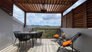 Ein überdachter Außenbereich mit Tisch und Stühlen, einem Liegestuhl und Blick auf die Landschaft.