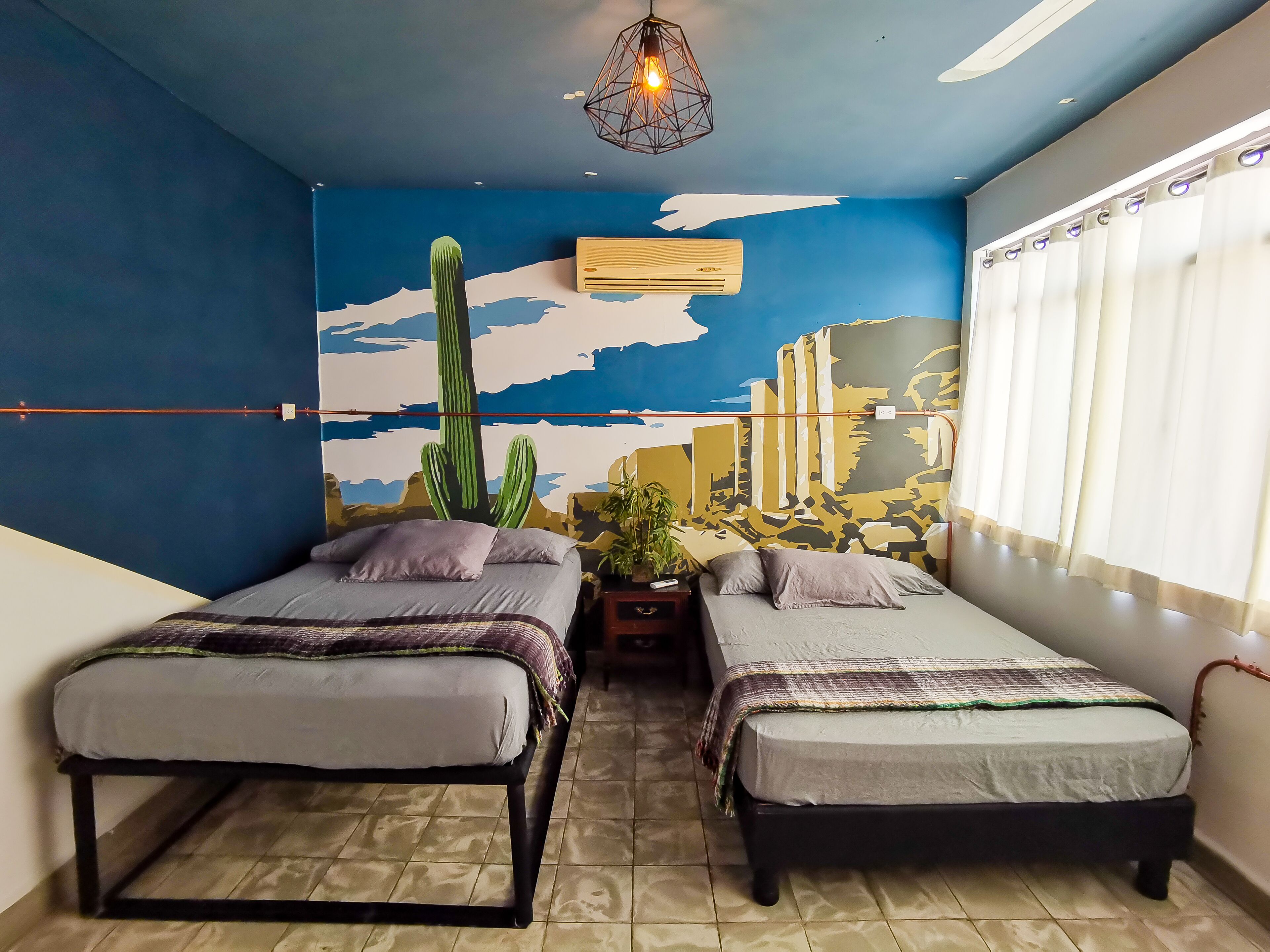 A room with two beds, a wall mural, an air conditioner, and a window with curtains.