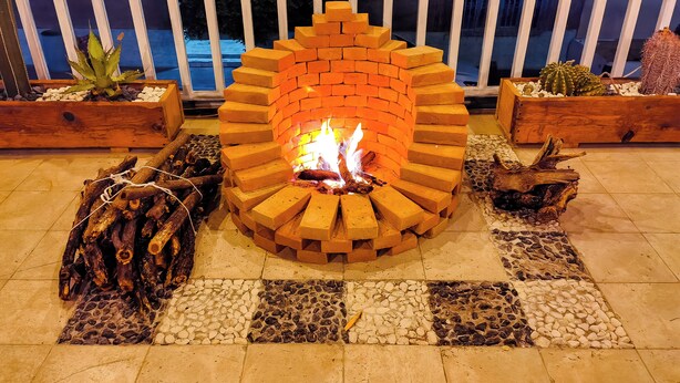 Een vuurkorf van baksteen met houtblokken en een brandend vuur, omringd door een betegeld terras en houten plantenbakken met cactussen.