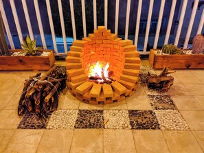 Ein Backstein-Feuerkorb mit Holzscheiten und brennendem Feuer, umgeben von einer gefliesten Terrasse und Holz-Pflanzkästen mit Kakteen.
