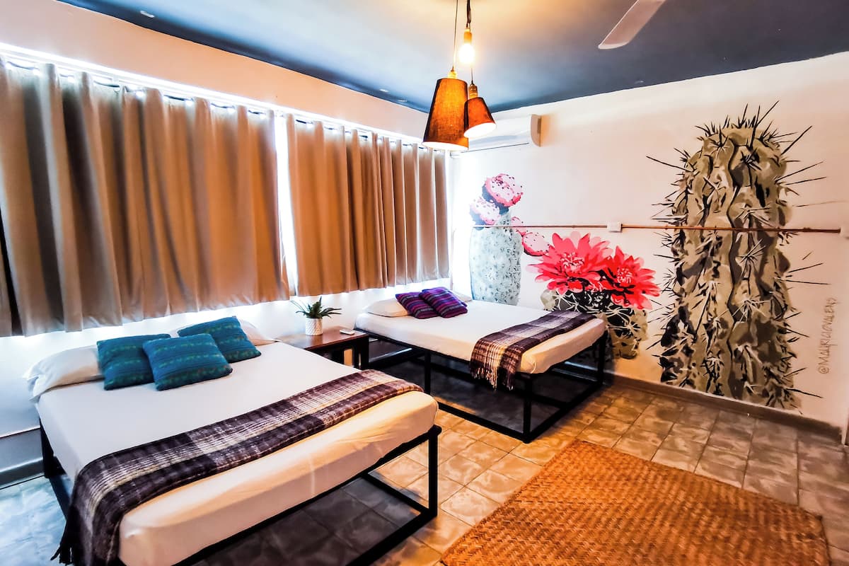 Habitación de hotel con dos camas, un ventilador de techo, un ventanal grande con cortinas, un cactus decorativo y una lámpara fijada a la pared.