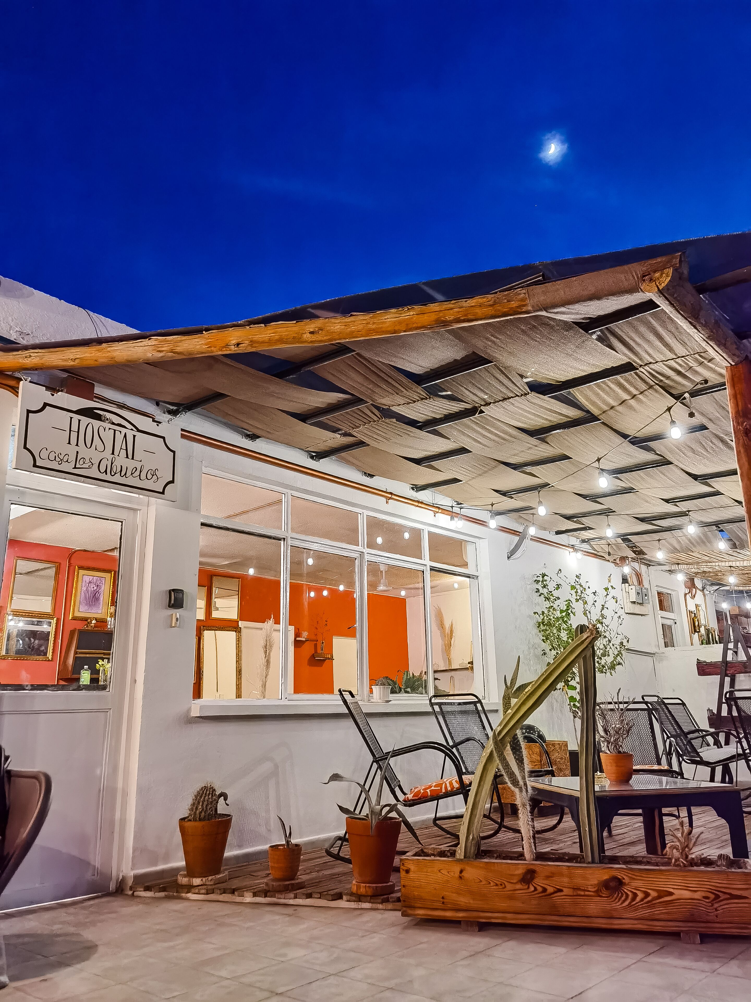 Un hostal acogedor llamado 'Hostal Casa los Abuelos', con un techo de madera rústica, ventanas amplias y diversas plantas en macetas.