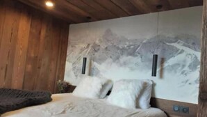 Un dormitorio con cama, almohadas, una colcha, un televisor montado en la pared y un cuadro grande de un paisaje de montaña.