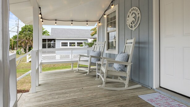 Eine überdachte Veranda mit Schaukelstühlen, einer weißen Tür und einer Fußmatte mit der Aufschrift 'Meet Me At The Beach'.