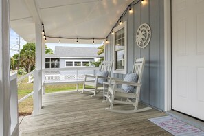 Un porche abrité avec des fauteuils à bascule, une porte blanche et un paillasson sur lequel est écrit : « Rendez-vous sur la plage ».