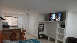 Een compacte kamer met een bed, een kleine kitchenette, een televisie en een zithoek met een stoel en een bankje.