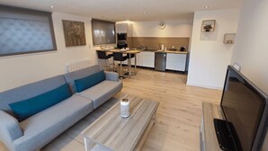 A modern living room with a grey sofa, a coffee table, and a TV. A kitchen with a sink and stove is visible in the background. - Welcome to the Gardens of Qiara.<br>Charming cottages in the heart of the Vosges Mo (Mattaincourt)