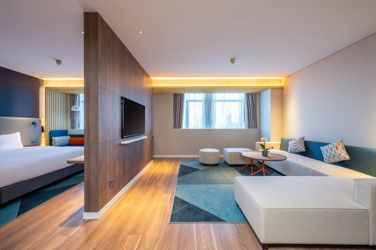 A modern hotel room with a bed, sofa, TV, and a small dining area.