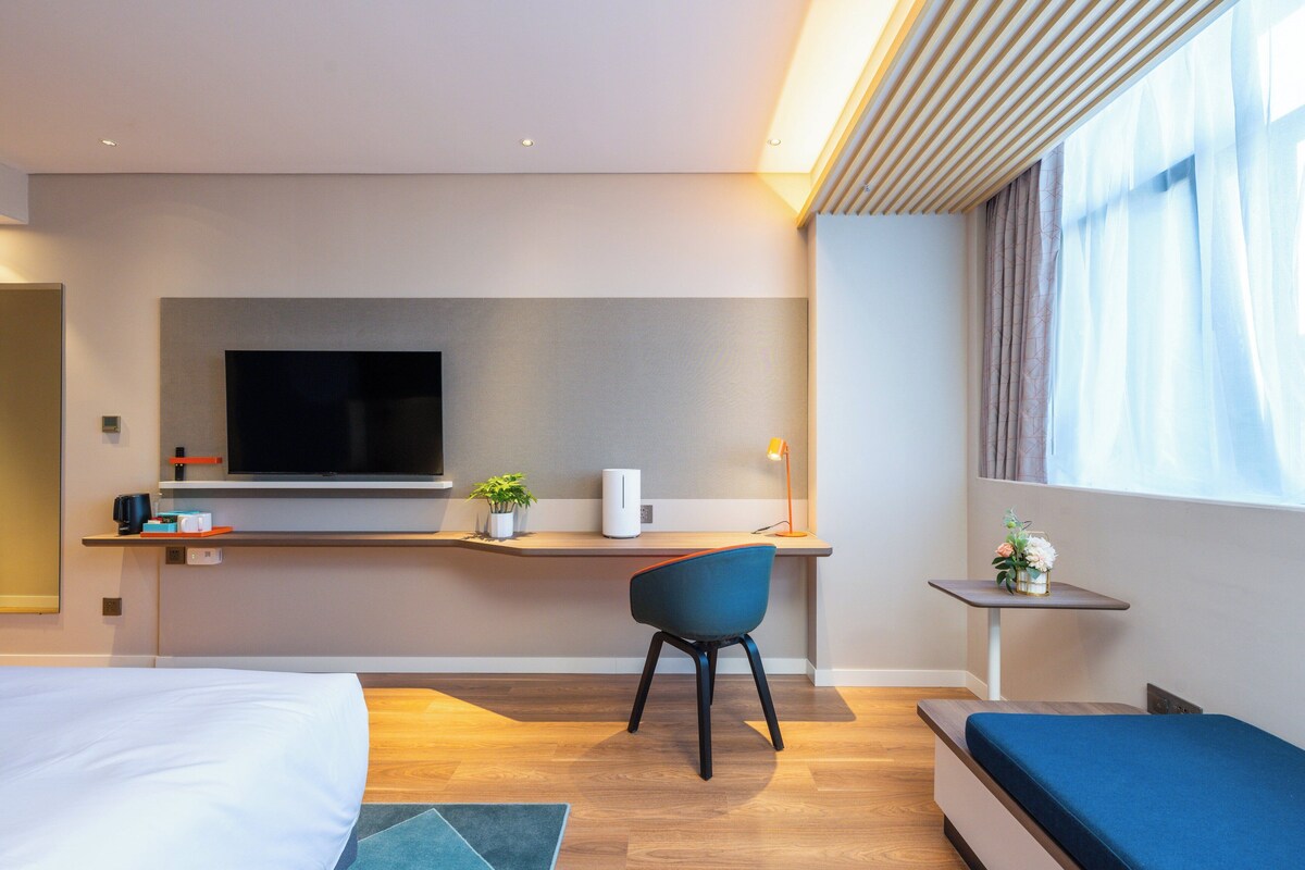 A modern hotel room with a bed, a desk with a flat-screen TV, a chair, a small table with a lamp, and a window with curtains.
