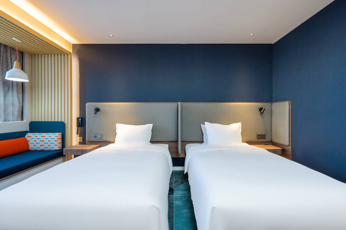 A hotel room with two single beds, a sofa with a patterned cushion, a wall-mounted telephone, and a ceiling light.