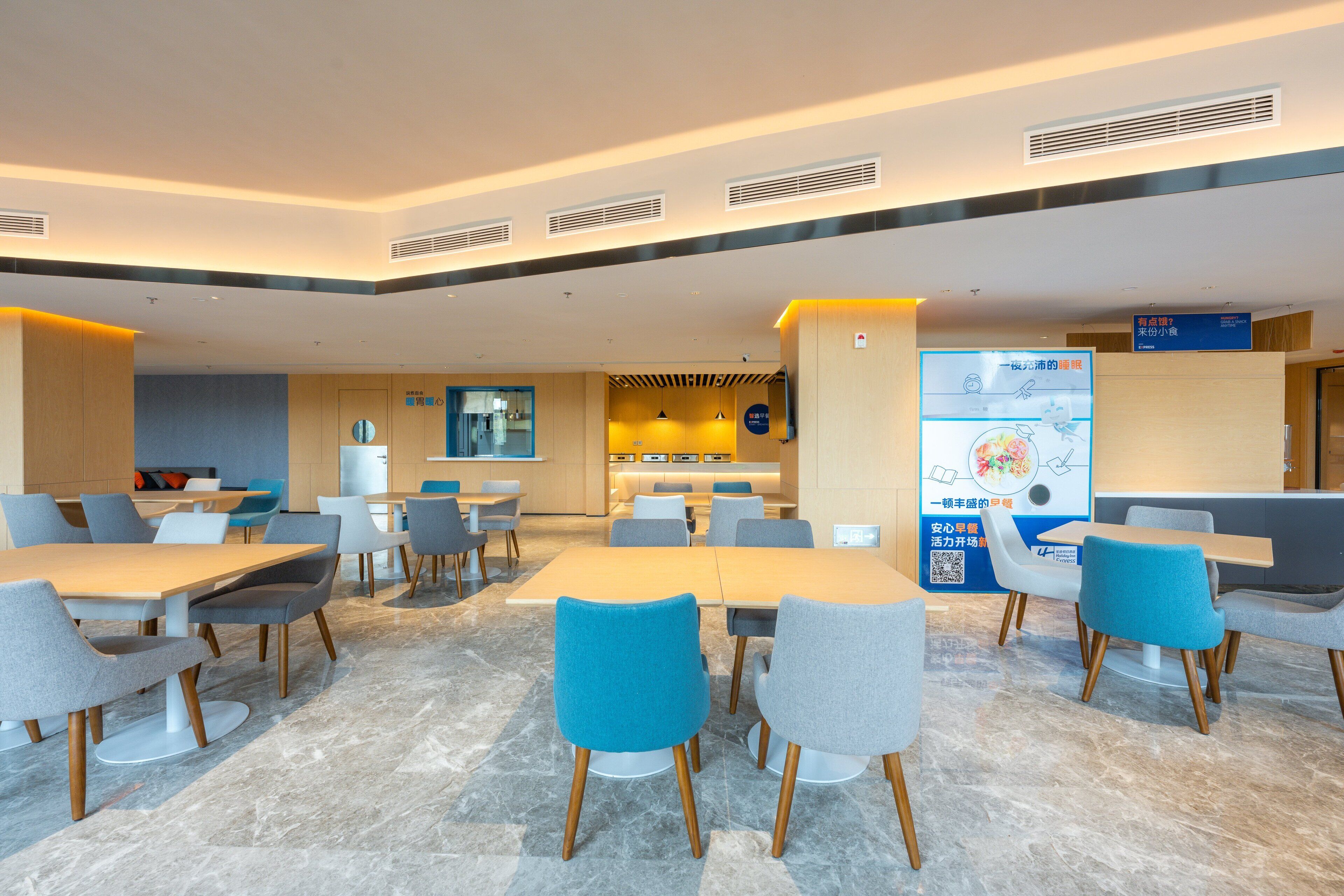 A modern hotel lobby with a central seating area, a kitchen in the background, and a digital information board.