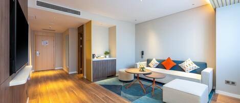 A modern living room with a sofa, ottoman, and coffee table, a kitchen area, and a flat-screen TV mounted on the wall.