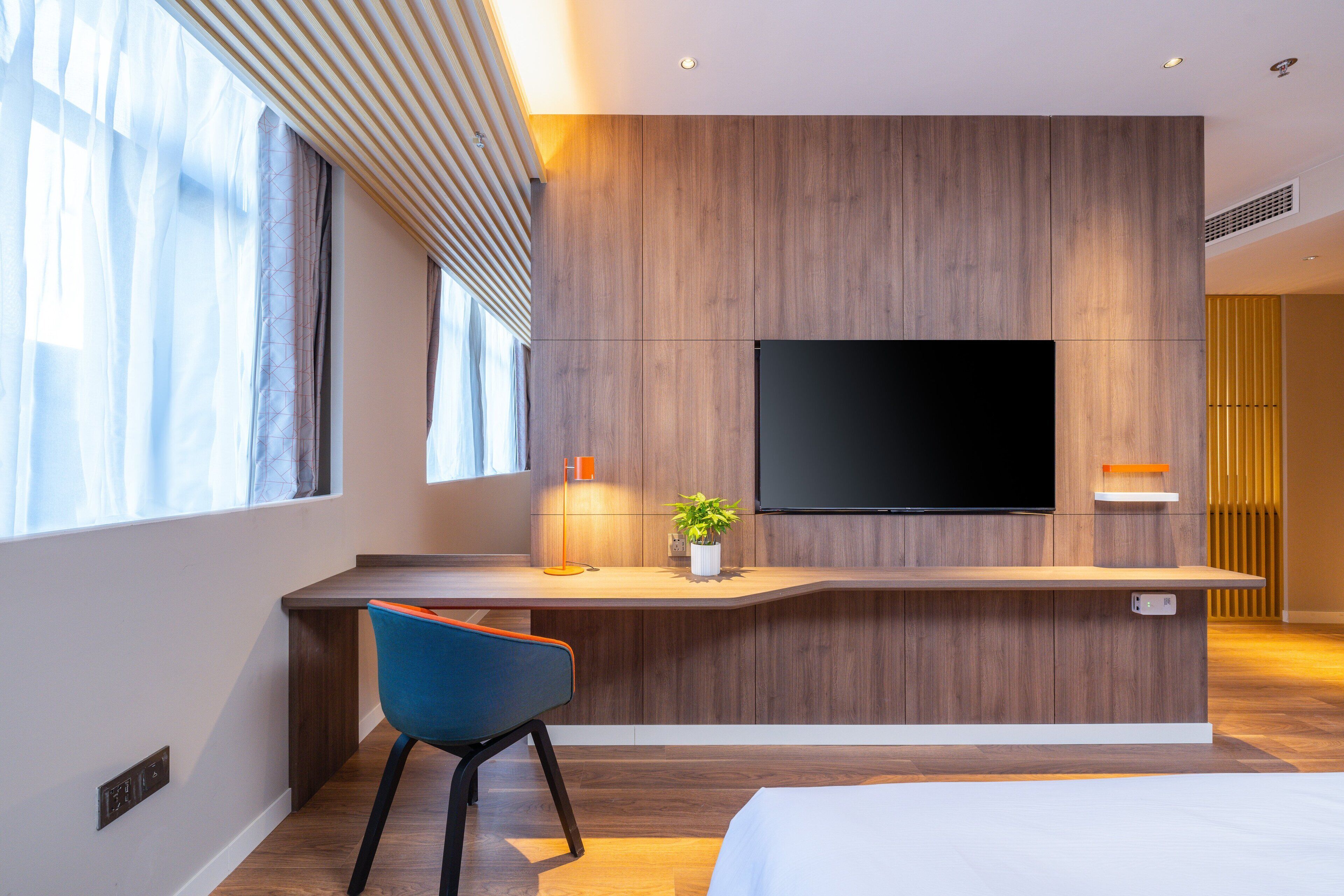 A modern hotel room with a wooden accent wall, a flat-screen TV mounted above a desk, a blue chair, and a bed with white bedding.