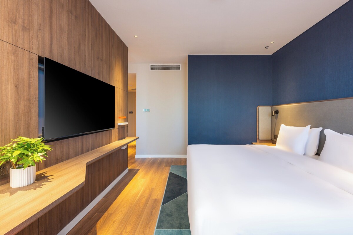 A modern hotel room with a large flat-screen TV mounted on a wooden panel, a bed with white linens, and a small potted plant on a wooden console table.
