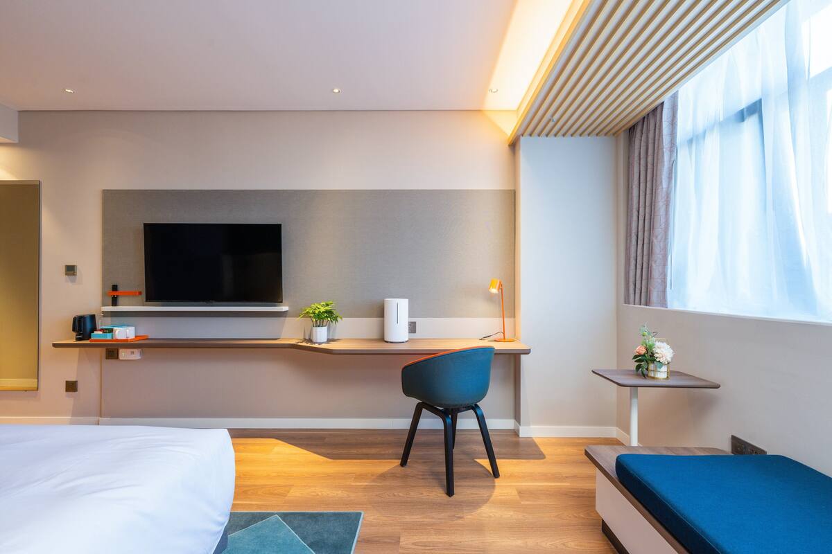 A modern hotel room with a bed, a desk with a flat-screen TV, a chair, a small table with a lamp, and a window with curtains.