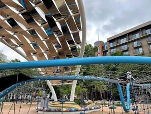 A playground with climbing structures and a large, modern building in the background. - Private Deck & Patio @Pearl/Riverwalk +Parking (San Antonio)