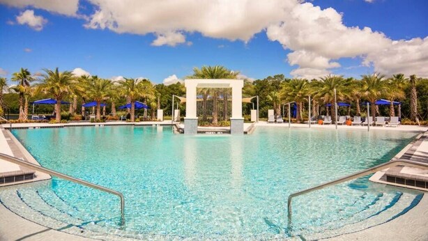 A clear blue pool with a central water feature, surrounded by palm trees and lounge chairs under blue umbrellas. - Relax in Style: Elegant Vacation Home in Orlando (Kissimmee)