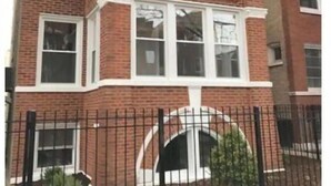Un edificio de ladrillo de dos pisos con un ventanal en el segundo piso, una cerca de metal negro en la fachada y un pequeño jardín con arbustos.