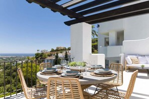 A terrace with a round table set for dining, wicker chairs, and a view of the landscape. - Altos La Quinta | Sea View Luxury Penthouse (Benahavís)