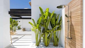A modern outdoor space with a white wall, a wooden door, and a shower area. There are large green plants and a seating area with cushions. - Altos La Quinta | Sea View Luxury Penthouse (Benahavís)