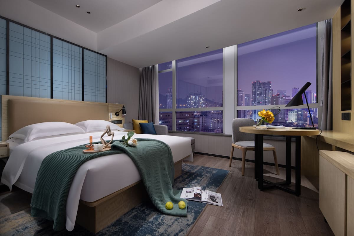 Una habitación de hotel moderna con una cama grande, un escritorio y vista a un paisaje urbano nocturno.