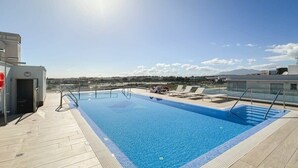 A rooftop swimming pool with clear blue water, a wooden deck, and a view of the cityscape in the background.