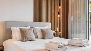 A neatly made bed with white linens and two folded towels, a wooden slat wall, and a glass door leading to a balcony.