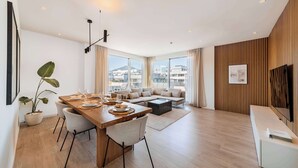 A modern living room with a wooden dining table, a sofa, and a large window offering a city view.