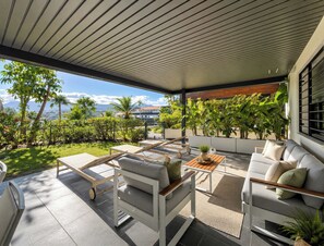 A covered outdoor seating area with a view of the ocean and mountains, furnished with white sofas and chairs, and surrounded by potted plants.