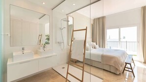 A modern bathroom with a large mirror, two sinks, a shower, and a bed with a view of the outdoors.