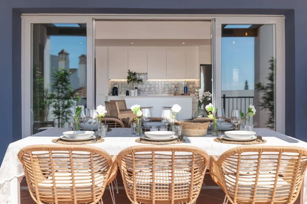Un espace de restauration en plein air avec une table dressée pour quatre personnes, entourée de chaises en osier, et une cuisine visible à travers une porte ouverte.