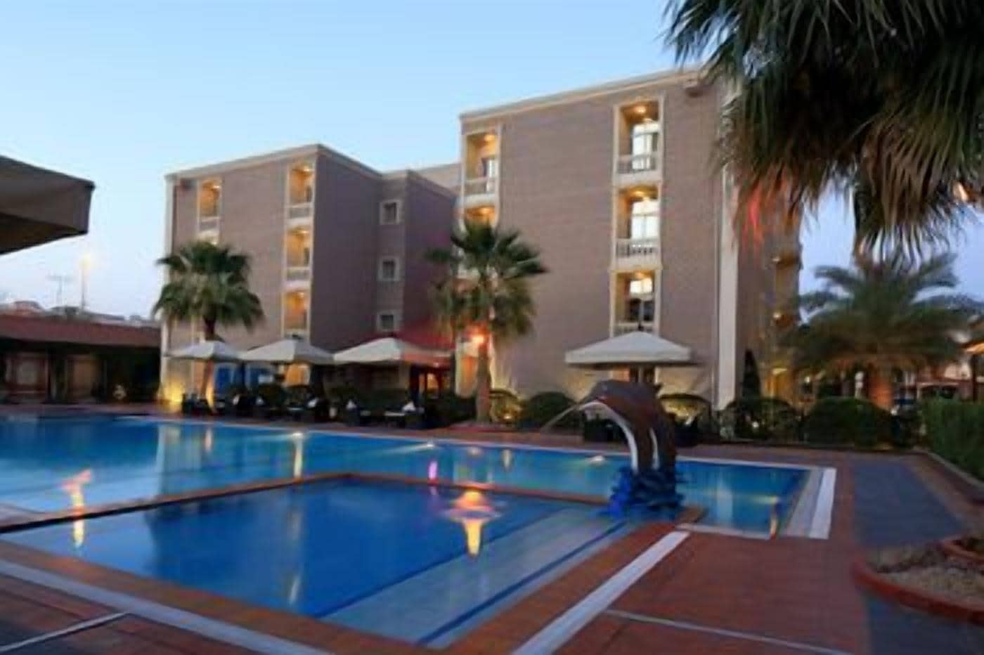 Ein Hotelpoolbereich mit rechteckigem Pool, Palmen und Sonnenschirmen.
