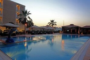 A swimming pool with lounge chairs and umbrellas, a building with multiple floors, and palm trees. - Boudl Gardenia (Al Khobar)