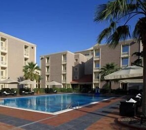 A swimming pool surrounded by multi-story buildings with balconies, palm trees, and umbrellas. - Boudl Gardenia (Al Khobar)