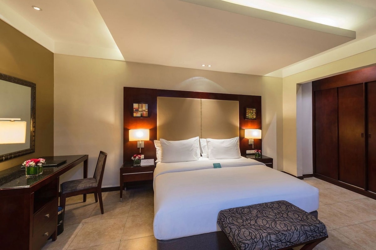 A hotel room with a bed, bedside lamps, a desk with a chair, a large mirror, and a wardrobe.