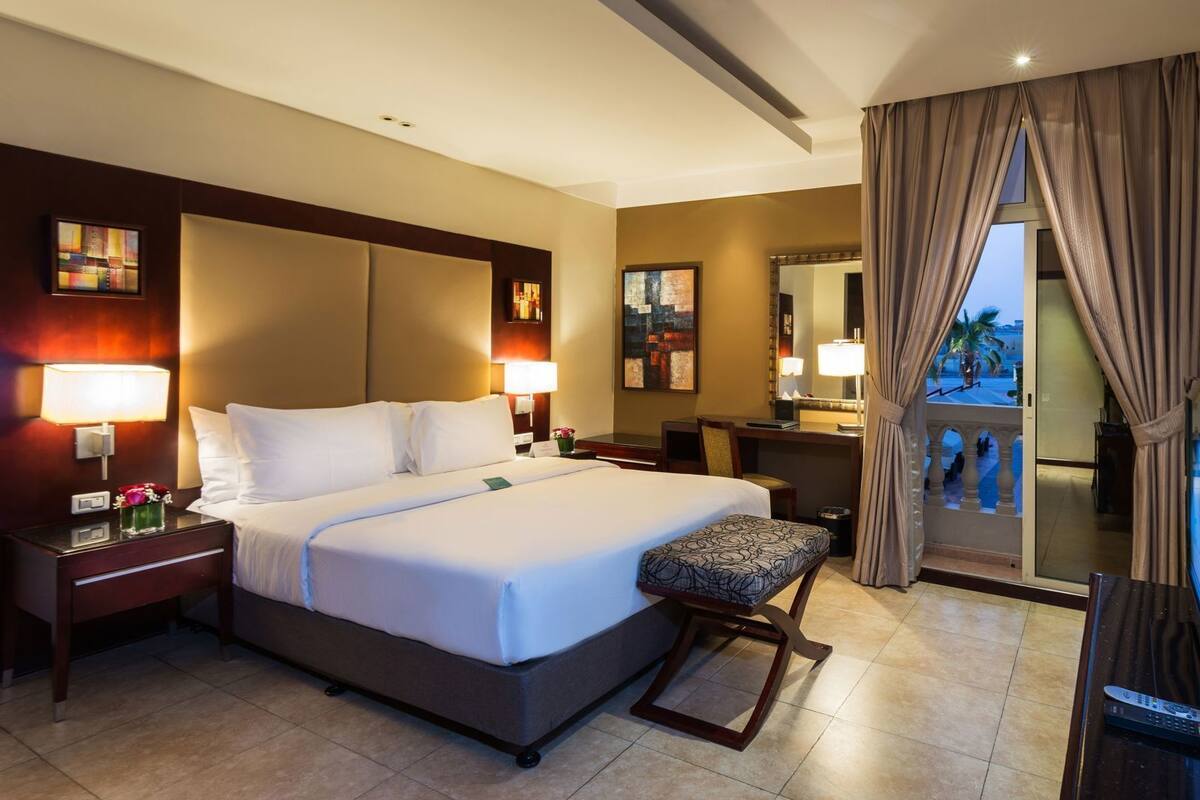 Une chambre d’hôtel comprenant un lit, des tables de chevet, un bureau, une chaise, une télévision et un balcon avec des rideaux.