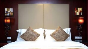 Une chambre d’hôtel avec un grand lit, deux coussins décoratifs, des appliques murales et une tête de lit de couleur neutre.