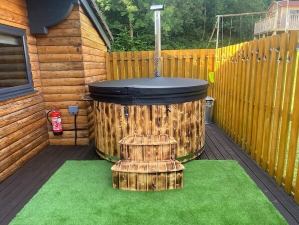 A hot tub with a wooden exterior, surrounded by a wooden deck and a fenced area. - The Cabin at Ffos Farm (Cefn cribwr)