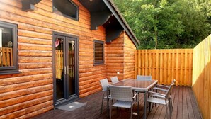 A wooden cabin with a deck, a glass door, and a table with chairs. - The Cabin at Ffos Farm (Cefn cribwr)