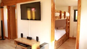 A room with a wooden bunk bed, a TV mounted on the wall, a wooden coffee table with speakers, and a radiator. - The Cabin at Ffos Farm (Cefn cribwr)