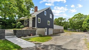 A two-story house with a dark exterior, a front porch, and a garage. - Cozy Cape Cod House - Stroll or Bike the Canal (Bourne)