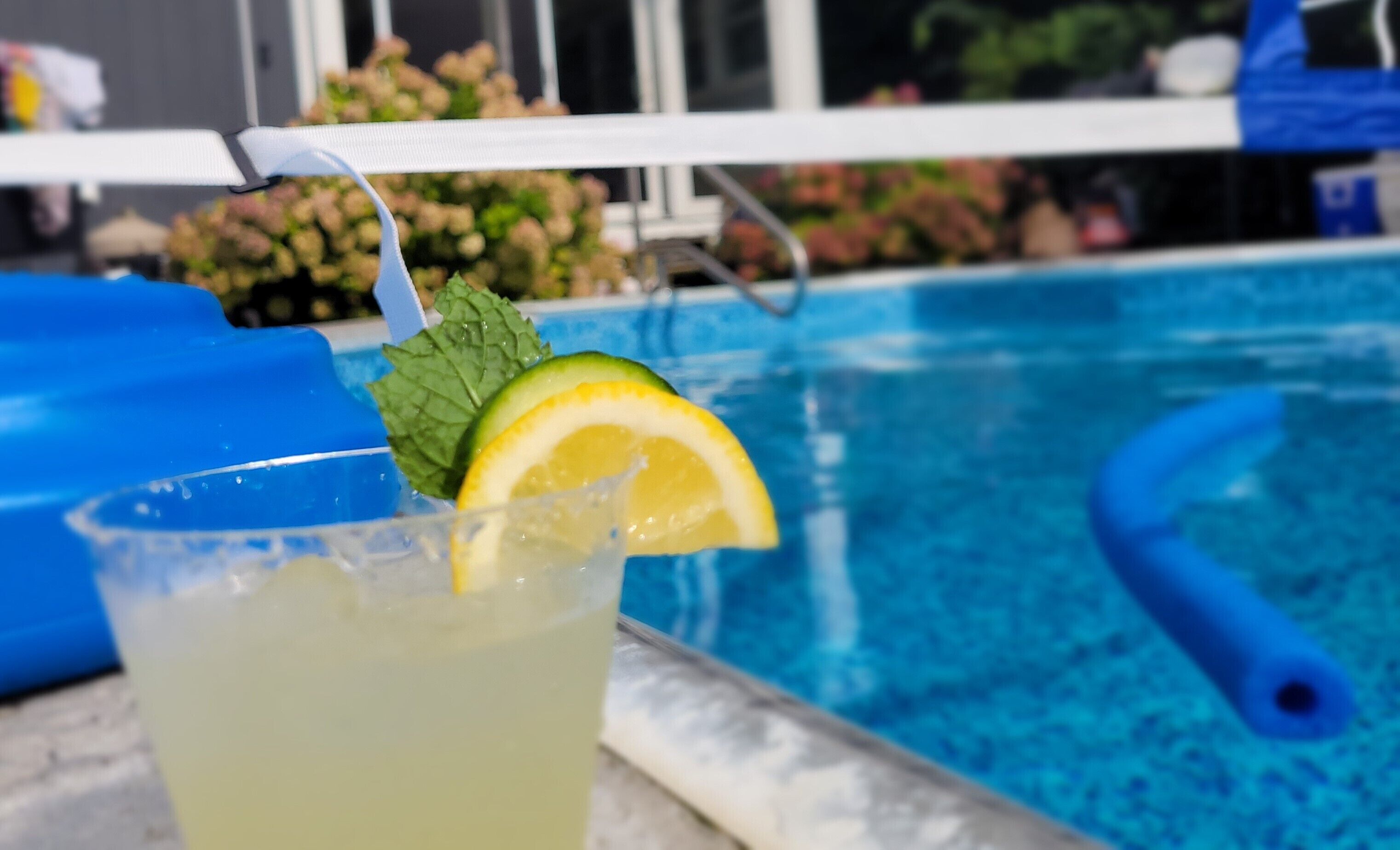 A poolside setting with a drink garnished with a lemon slice and mint, a blue pool float, and a pool in the background.