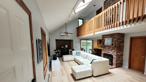 A spacious living area with a sectional sofa, a fireplace, and a balcony with wooden railings. - Cozy Cape Cod House - Stroll or Bike the Canal (Bourne)