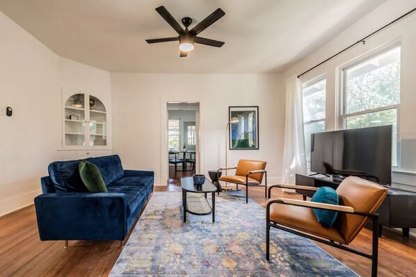 A spacious living room with a blue sofa, a brown leather chair, a coffee table, and a TV. There is a ceiling fan, a bookshelf, and a window with a view. - Private Deck & Patio with Parking- 3blks to Pearl food & fun! (San Antonio)