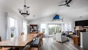 Uma sala de estar espaçosa com uma grande mesa de jantar de madeira, assentos confortáveis, um ventilador de teto e uma estante de livros.