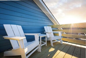 A covered outdoor seating area with white Adirondack chairs and a small table, set against a blue wooden wall.
