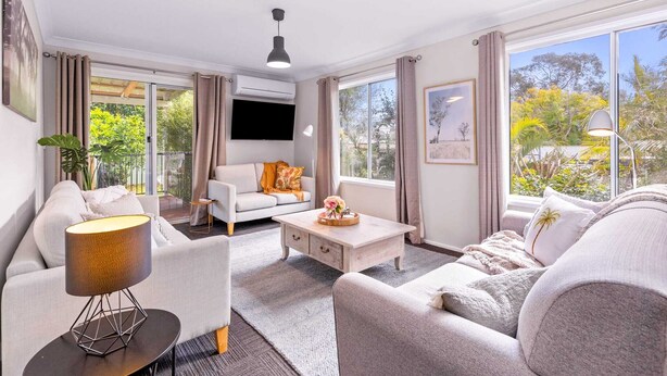 A modern living room with a sofa, a coffee table, and a lamp. There is a TV mounted on the wall and a window with curtains.