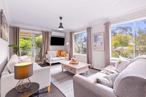 Ein modernes Wohnzimmer mit einer Couch, einem Couchtisch und einer Lampe. An der Wand ist ein Fernseher befestigt und es gibt ein Fenster mit Vorhängen.