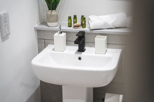 A bathroom sink with a white basin, a black faucet, and two soap dispensers. There is a potted plant and a towel rack with a white towel above the sink. - Birtin Works Apartments - Brand New - City Centre (Sheffield)