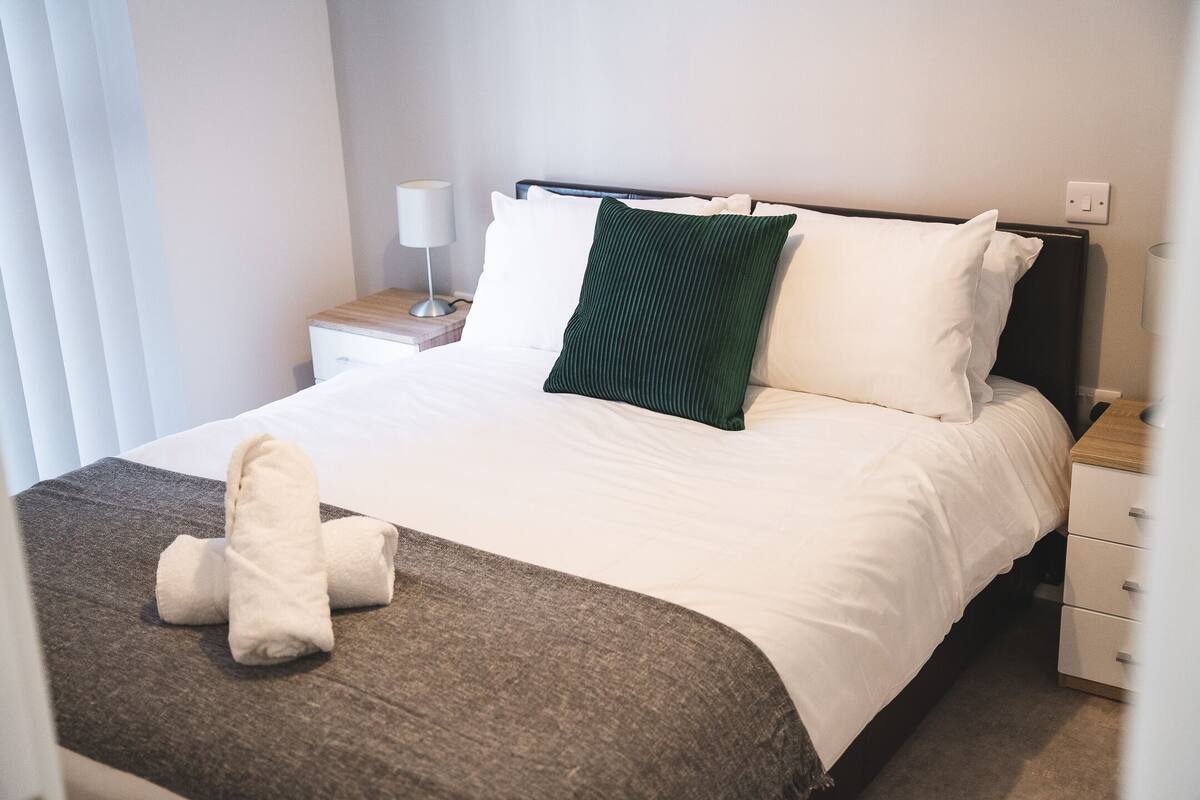 A neatly made bed with white and green pillows, a brown throw blanket, and a pair of folded towels on the bed.